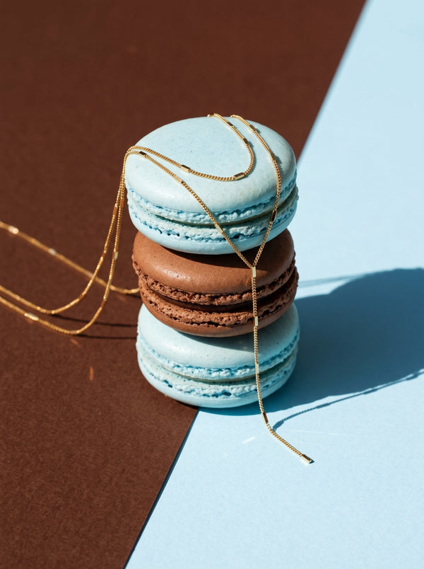Lundholm Nathalie layered ketting met Y-hanger goudkleurig over een stapel blauwe en bruine macarons