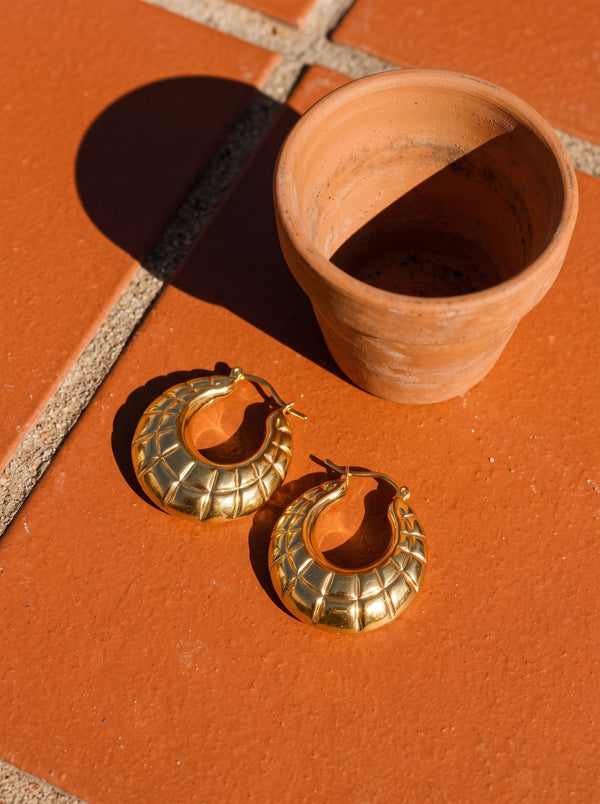 Lundholm Skye grote oorringen met reliëf goudkleurig op warme terracotta tegels naast een bloempot