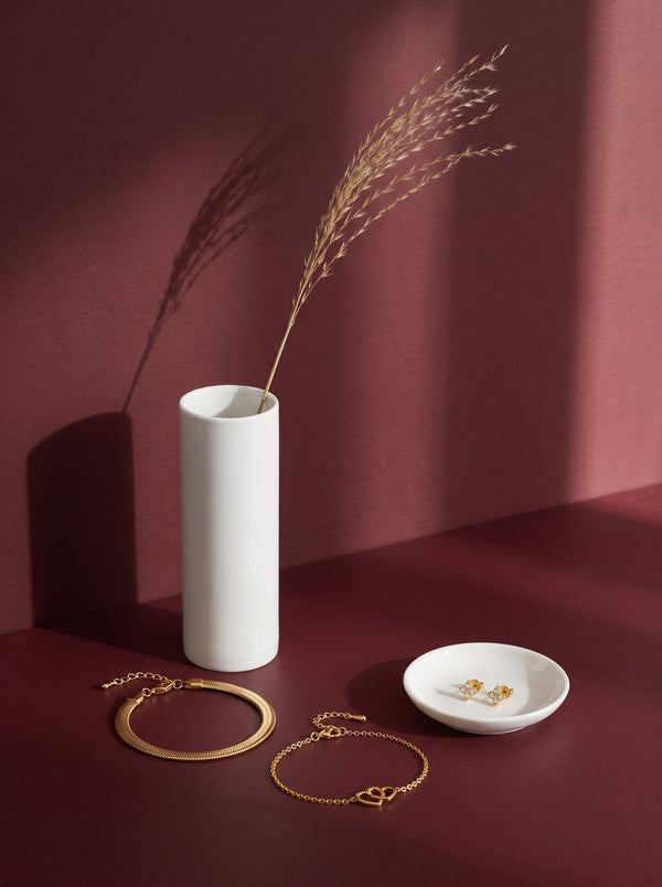 Lundholm Danielle snake armband, Alice armband met hartjes en Livan oorknopjes goudkleurig bij een witte vaas met pluimgras tegen bordeaux en oudroze