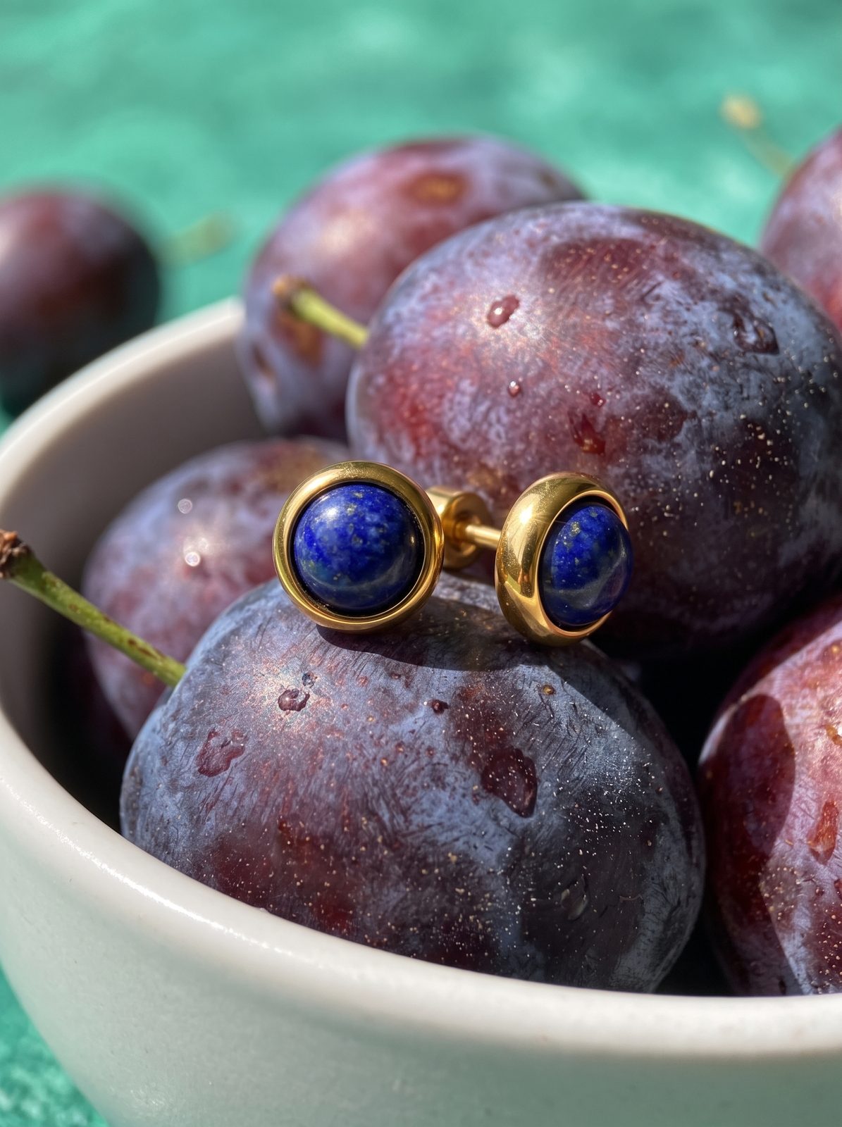 Goudkleurige oorbellen met blauwe steen op een schaal pruimen