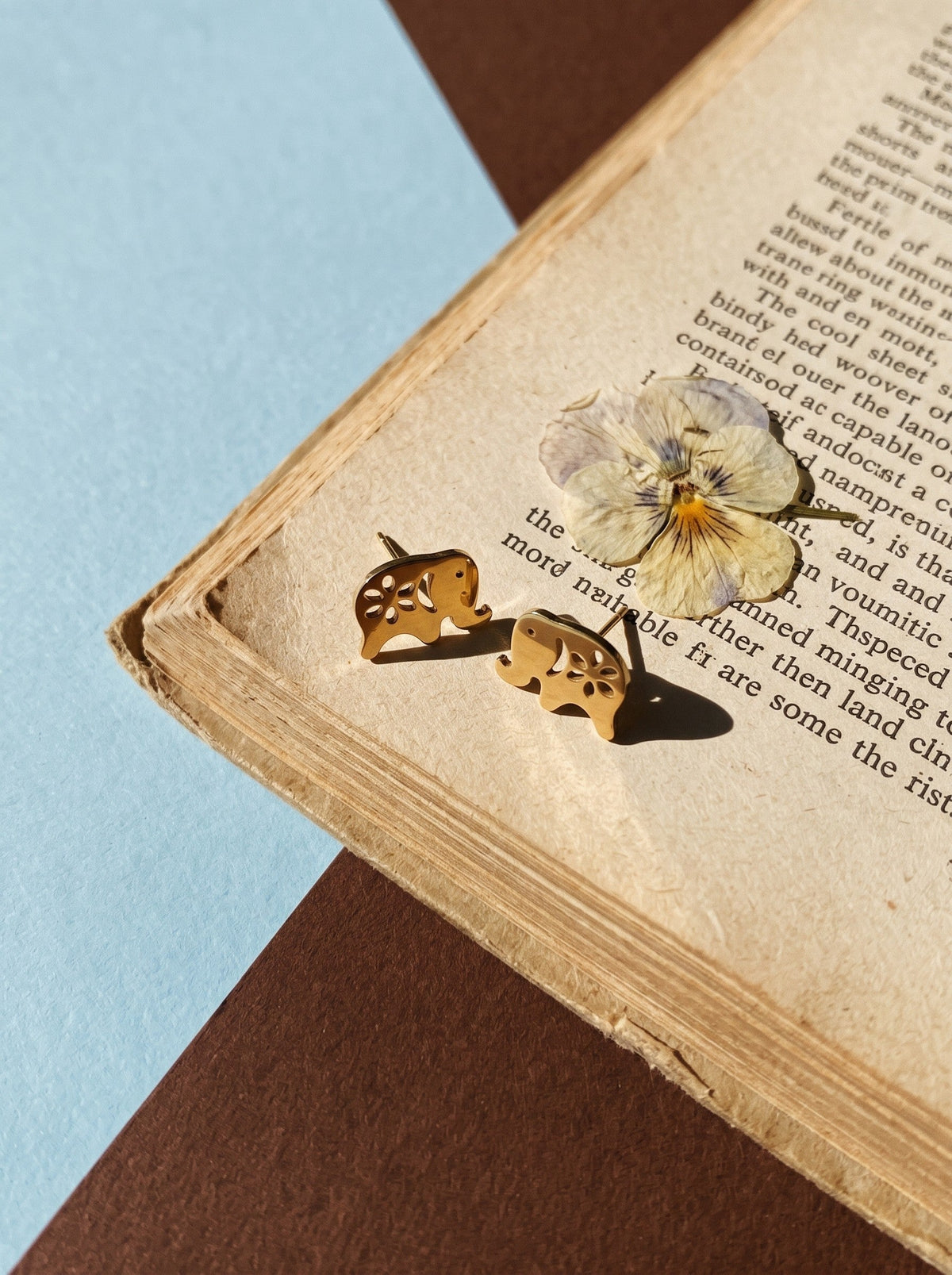 Lundholm Lucky olifantjes oorknopjes goudkleurig op een oud boek met een gedroogd viooltje