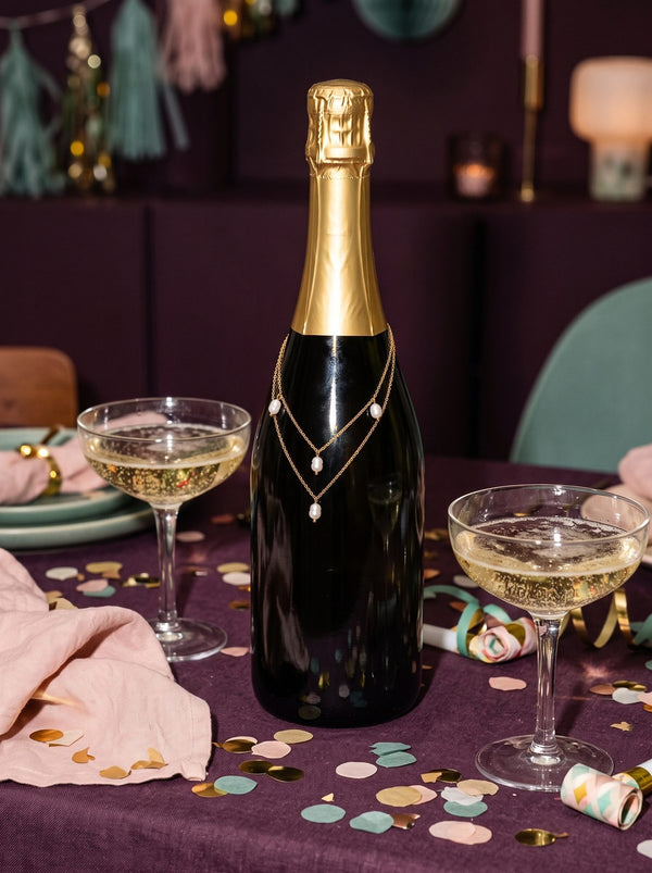 Lundholm Kristy dubbellaagse parelketting goudkleurig om een fles champagne tussen coupes en confetti op een pruimkleurige tafel