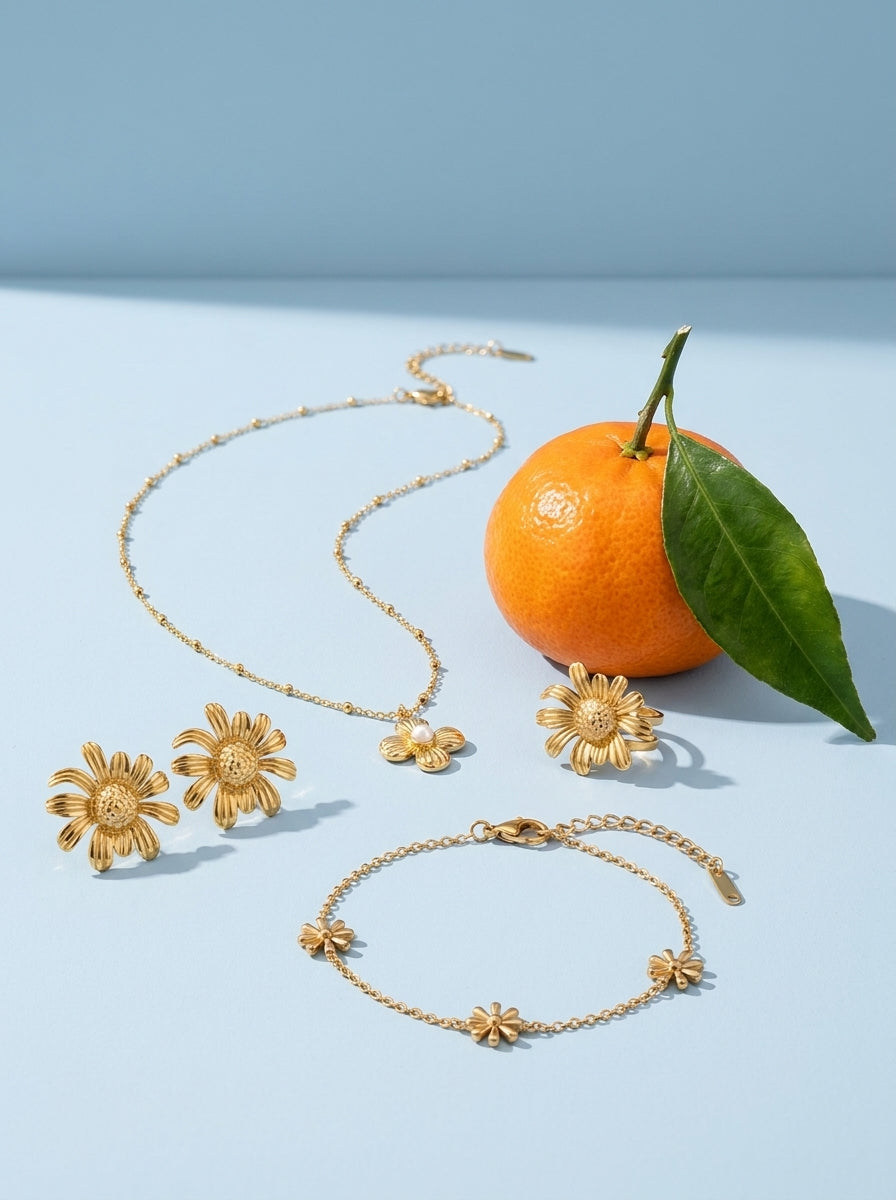 Lundholm Äng look in gold tone: Imogen necklace with a little flower, Marguerite daisy earrings, Smilla daisy bracelet and Marguerite daisy ring by a mandarin against light blue