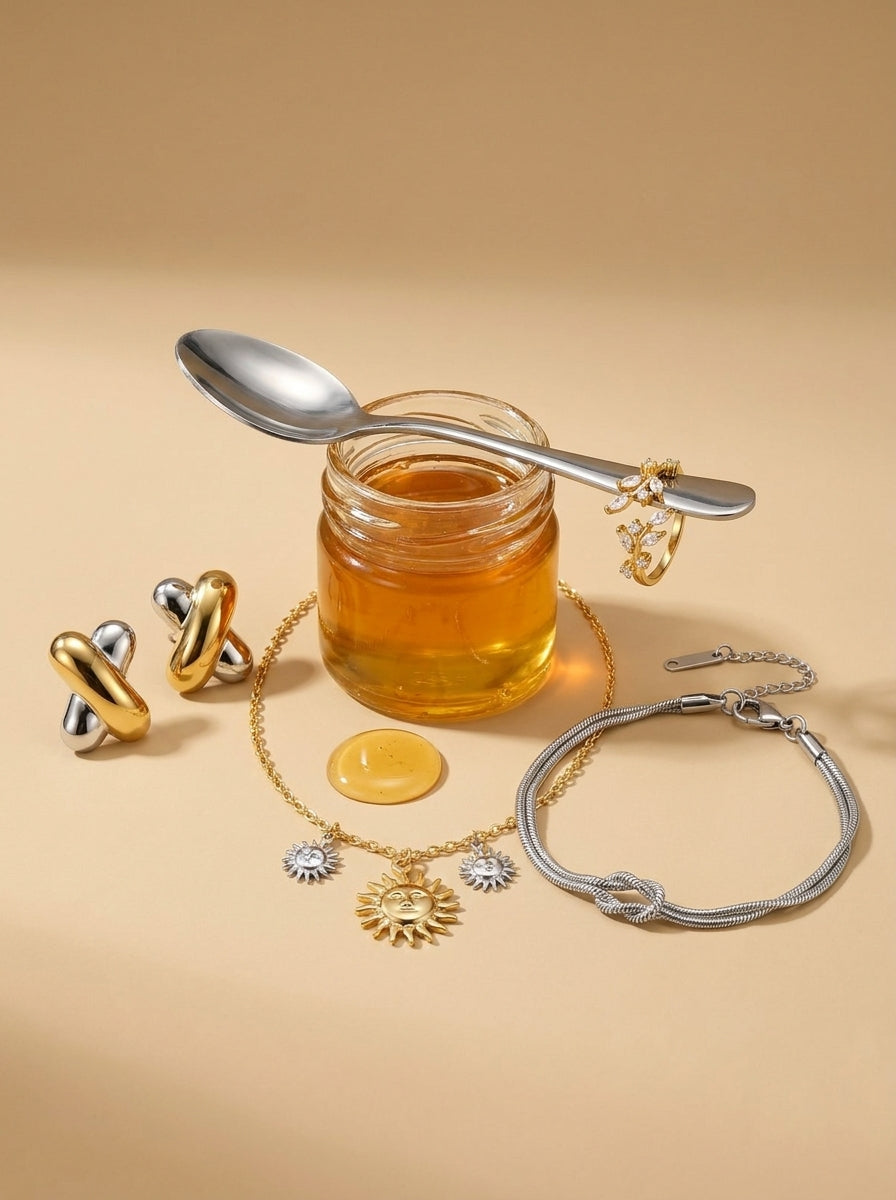 Lundholm Blanda look in gold and silver: Helia necklace with little suns, Hera two-tone studs, Brianna bracelet with a knot and Sanna leaf ring by a jar of honey against warm sand