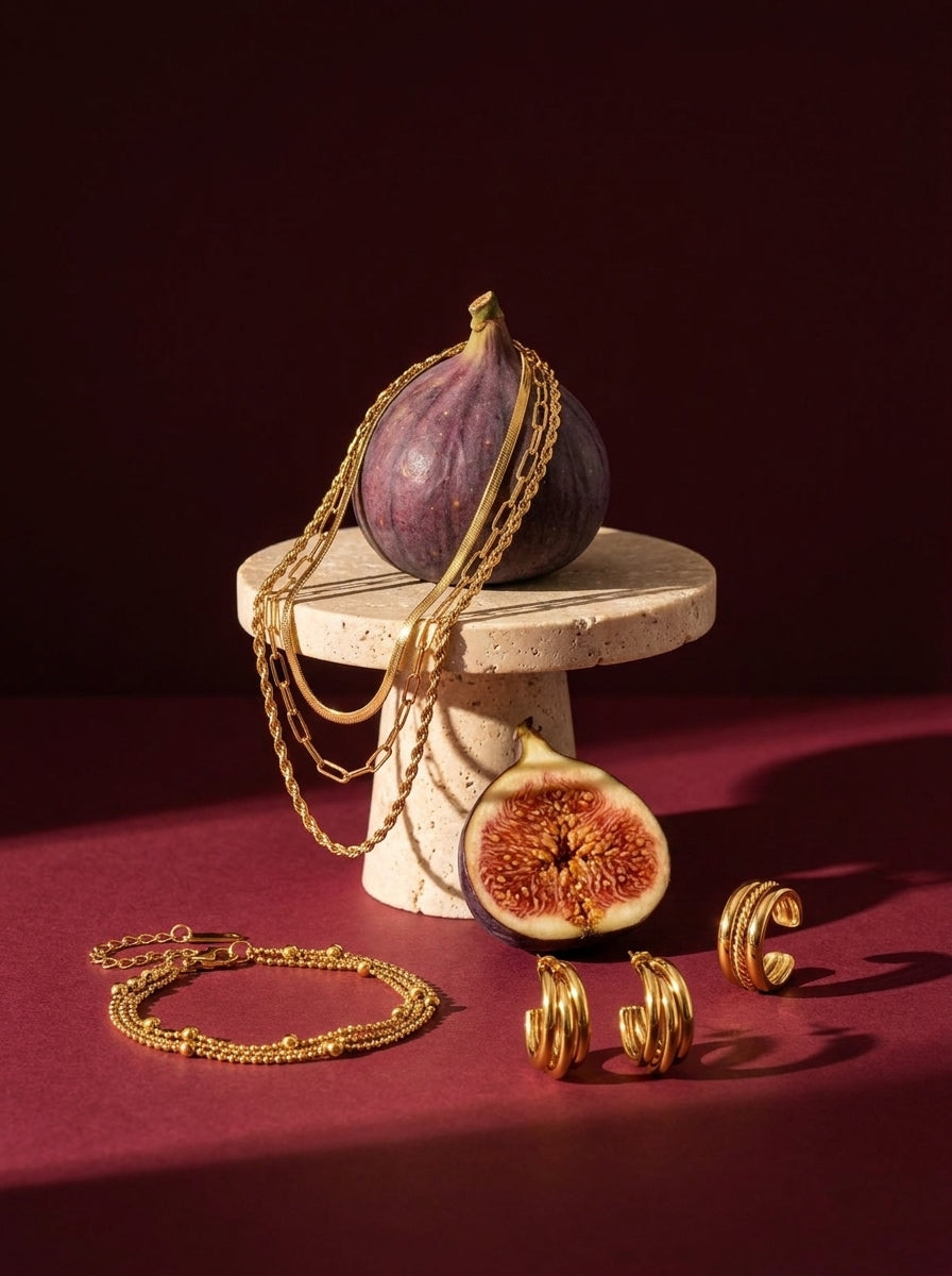 Lundholm Look Tre in Goldfarbe: Manou dreilagige Kette, Nika Triple-Hoop-Ohrringe, Alina dreireihiges Armband und Billie Ring neben frischen Feigen vor Bordeaux