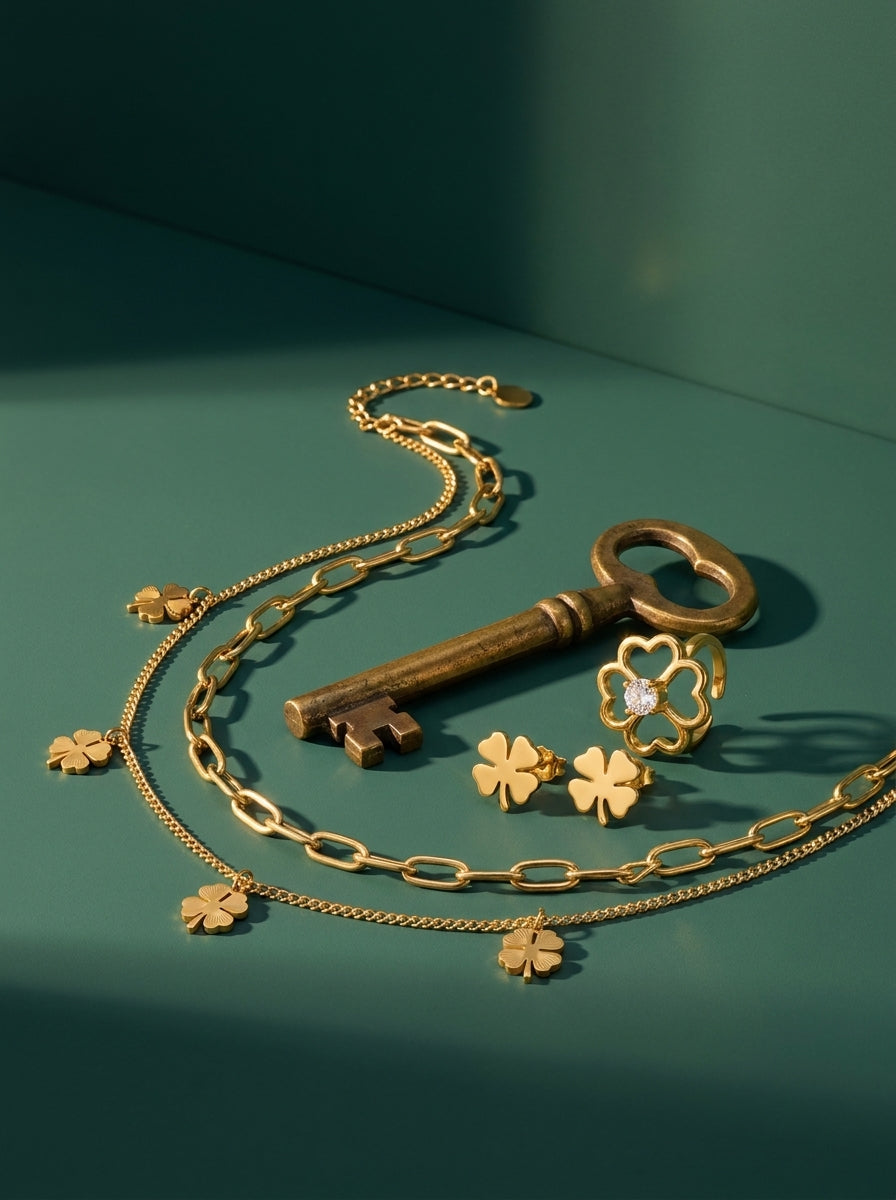 Lundholm look Tur in goudkleurig: Georgia layered klavertjevier ketting, Klara klavertjevier oorknopjes en Bloom ring met hartjes naast een oude sleutel tegen zeegroen