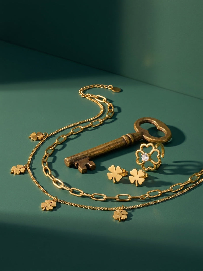 Lundholm look Tur in goudkleurig: Georgia layered klavertjevier ketting, Klara klavertjevier oorknopjes en Bloom ring met hartjes naast een oude sleutel tegen zeegroen