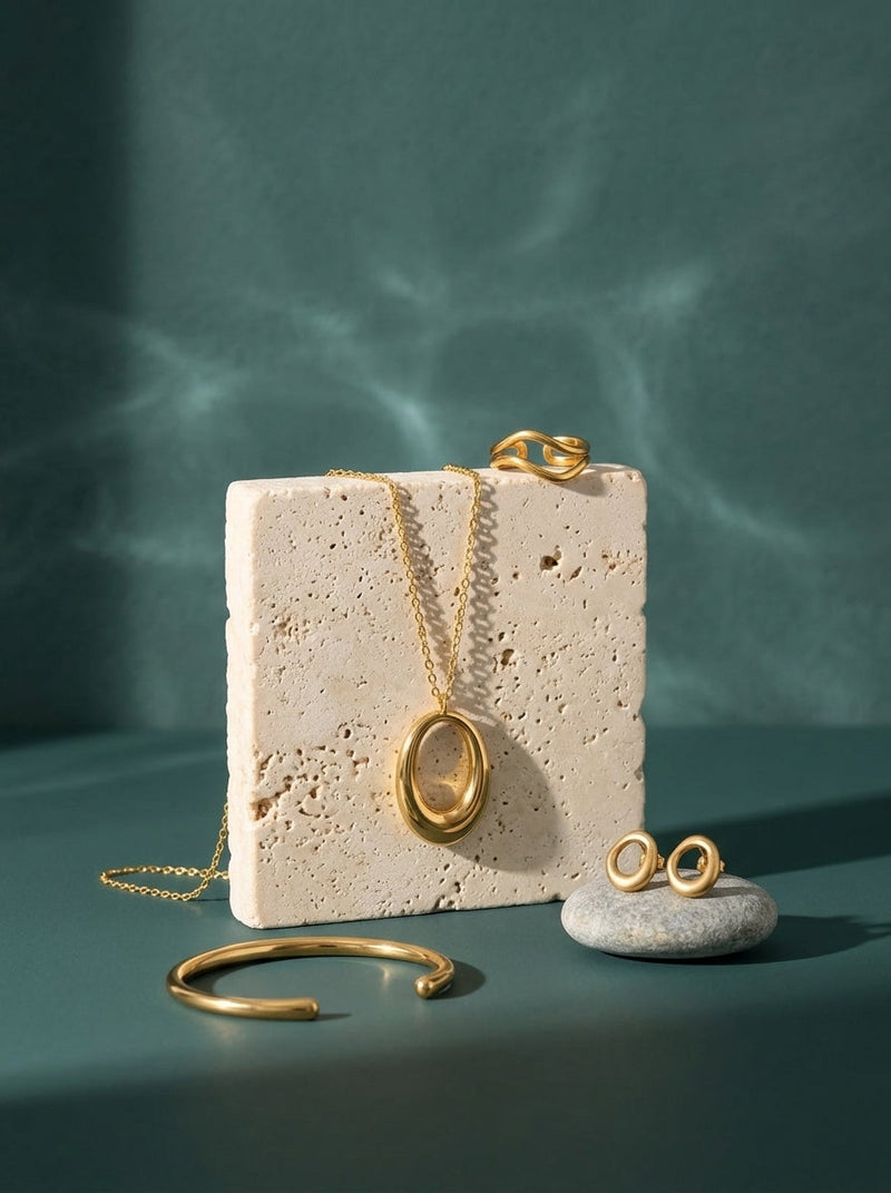 Lundholm look Våg in goudkleurig: Ava lange ketting, Oona oorknopjes, Runa bangle en Shelby ring op travertin tegen zeegroen