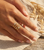 Lundholm Alexia goudkleurige verstelbare spiraal ring voor dames met drie fonkelende zirkonia steentjes, gedragen door vrouw met hand op linnen en tarwe halmen voor warme zomerse lookbook lifestyle shoot. Premium stainless steel met PVD gold plating van 2.5 micron, waterproof en hypoallergeen. Een verfijnd cadeau voor vrouw, moeder of vriendin op Moederdag of verjaardag in Scandinavisch design.