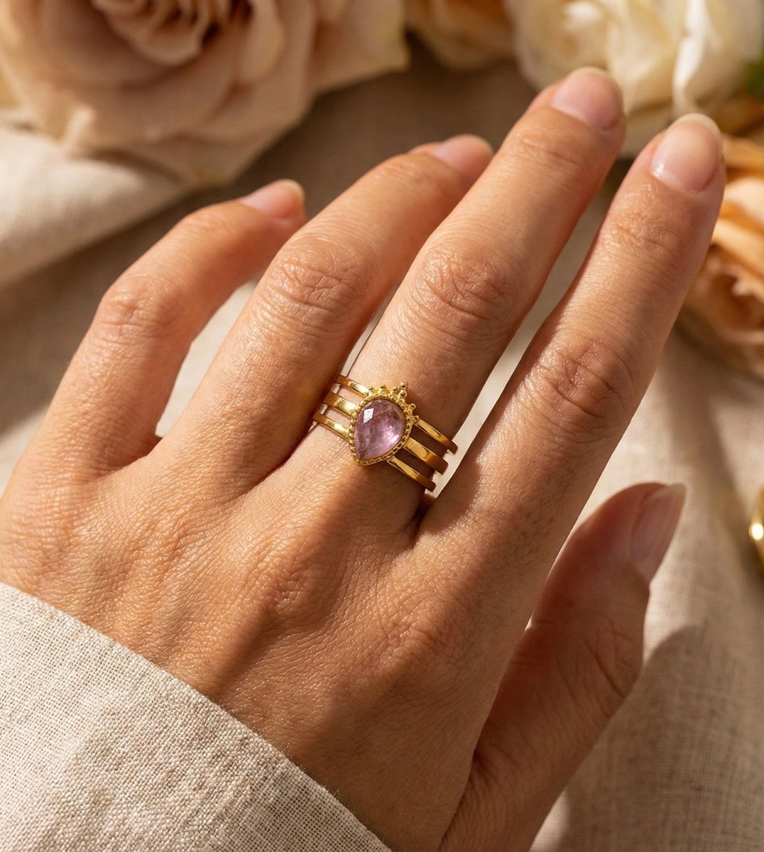 Lundholm Amara Serie luxe ring voor dames in goudkleurig met paarse steen op drie laags band gedragen aan hand van model in lifestyle setting met rozen, gemaakt van premium stainless steel met PVD coating van 2.5 micron, waterproof en hypoallergeen, perfect cadeau voor vrouw, moederdag of verjaardag in Scandinavisch design.