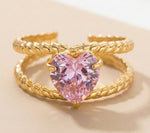 Lundholm Francine goudkleurige verstelbare statement ring voor dames met groot roze hart steen op dubbele getwist touw band, gefotografeerd op crème ondergrond voor romantische productshot. Premium stainless steel met PVD gold plating van 2.5 micron, waterproof en hypoallergeen. Een romantisch cadeau voor vrouw, moeder of vriendin op Moederdag, Valentijnsdag of verjaardag in Scandinavisch design.