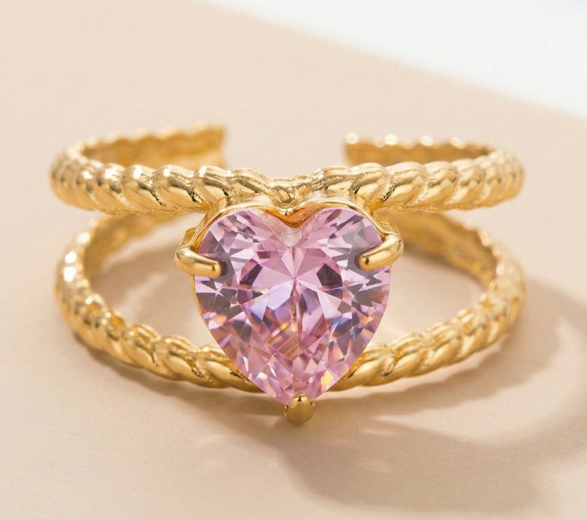 Lundholm Francine goudkleurige verstelbare statement ring voor dames met groot roze hart steen op dubbele getwist touw band, gefotografeerd op crème ondergrond voor romantische productshot. Premium stainless steel met PVD gold plating van 2.5 micron, waterproof en hypoallergeen. Een romantisch cadeau voor vrouw, moeder of vriendin op Moederdag, Valentijnsdag of verjaardag in Scandinavisch design.
