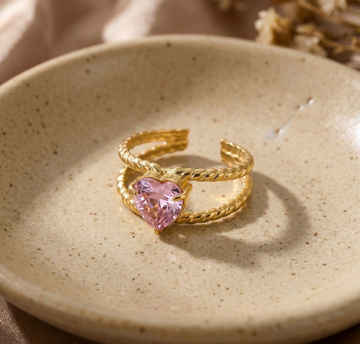 Lundholm Francine goudkleurige verstelbare statement ring voor dames met groot roze hart steen op getwist touw band, gefotografeerd op crème keramische schaal voor warme productshot. Premium stainless steel met PVD gold plating van 2.5 micron, waterproof en hypoallergeen. Een romantisch cadeau voor vrouw, moeder of vriendin op Moederdag, Valentijnsdag of verjaardag in Scandinavisch design.
