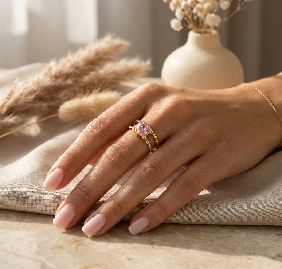 Lundholm Francine goudkleurige verstelbare statement ring voor dames met roze hart steen, gedragen door vrouw met hand op linnen en marmer met droogbloemen vaas voor warme lookbook lifestyle shoot. Premium stainless steel met PVD gold plating van 2.5 micron, waterproof en hypoallergeen. Een romantisch cadeau voor vrouw, moeder of vriendin op Moederdag, Valentijnsdag of verjaardag in Scandinavisch design.