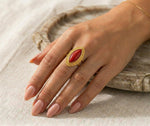 Lundholm Ivana goudkleurige verstelbare statement ring voor dames met grote rode ovale steen in vintage ontwerp, gedragen door vrouw met hand op linnen en rotan voor warme lookbook lifestyle shoot. Premium stainless steel met PVD gold plating van 2.5 micron, waterproof en hypoallergeen. Een opvallend cadeau voor vrouw, moeder of vriendin op Moederdag of verjaardag in Scandinavisch design.