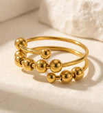 Lundholm Lauren goudkleurige verstelbare anxiety ring voor dames met schuifbare bolletjes en kralen voor stress relief en fidget, gefotografeerd op witte natuursteen voor stijlvolle productshot. Premium stainless steel met PVD gold plating van 2.5 micron, waterproof en hypoallergeen. Een doordacht cadeau voor vrouw, moeder of vriendin op Moederdag of verjaardag in Scandinavisch design.