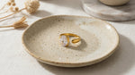 Lundholm Lenore goudkleurige verstelbare open statement ring voor dames met grote ovale zirkonia steen, gefotografeerd op crème keramisch schaaltje met droogbloemen voor warme productshot. Premium stainless steel met PVD gold plating van 2.5 micron, waterproof en hypoallergeen. Een opvallend cadeau voor vrouw, moeder of vriendin op Moederdag of verjaardag in Scandinavisch design.