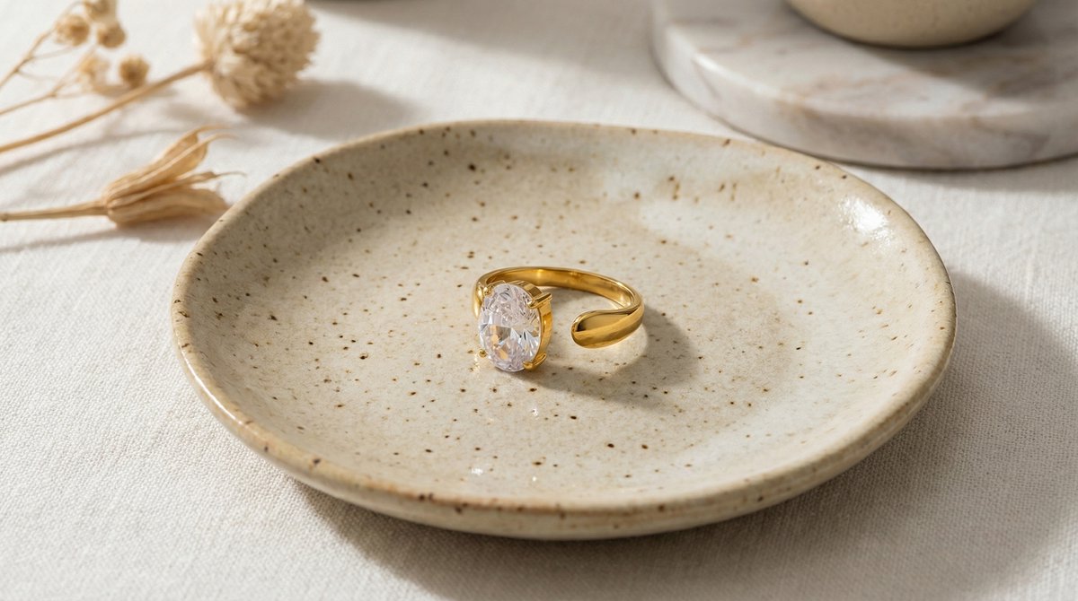 Lundholm Lenore goudkleurige verstelbare open statement ring voor dames met grote ovale zirkonia steen, gefotografeerd op crème keramisch schaaltje met droogbloemen voor warme productshot. Premium stainless steel met PVD gold plating van 2.5 micron, waterproof en hypoallergeen. Een opvallend cadeau voor vrouw, moeder of vriendin op Moederdag of verjaardag in Scandinavisch design.