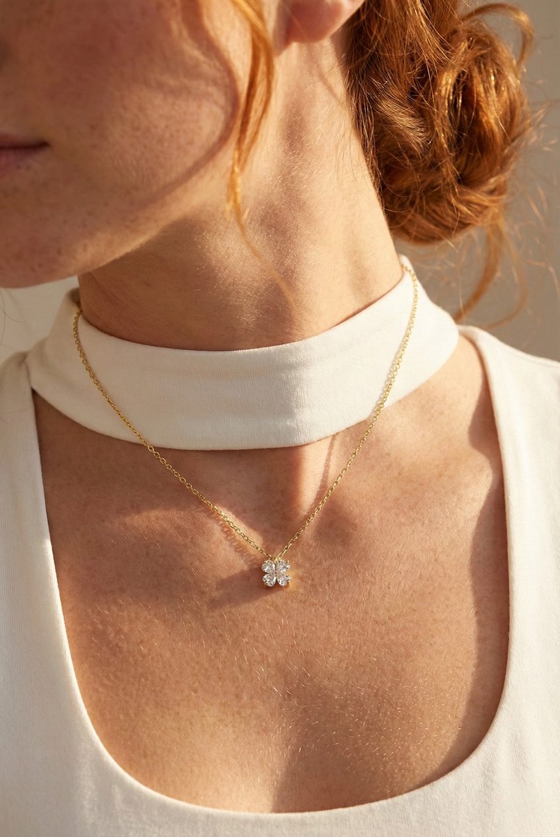 Lundholm Lukk goudkleurige geluksketting voor dames met klavertje vier hanger ingelegd met zirkonia, gedragen door vrouw met witte top en sjaal voor lookbook lifestyle shoot. Premium stainless steel met PVD gold plating van 2.5 micron, waterproof en hypoallergeen. Een betekenisvol cadeau voor vrouw, moeder of vriendin op Moederdag of verjaardag in Scandinavisch design.