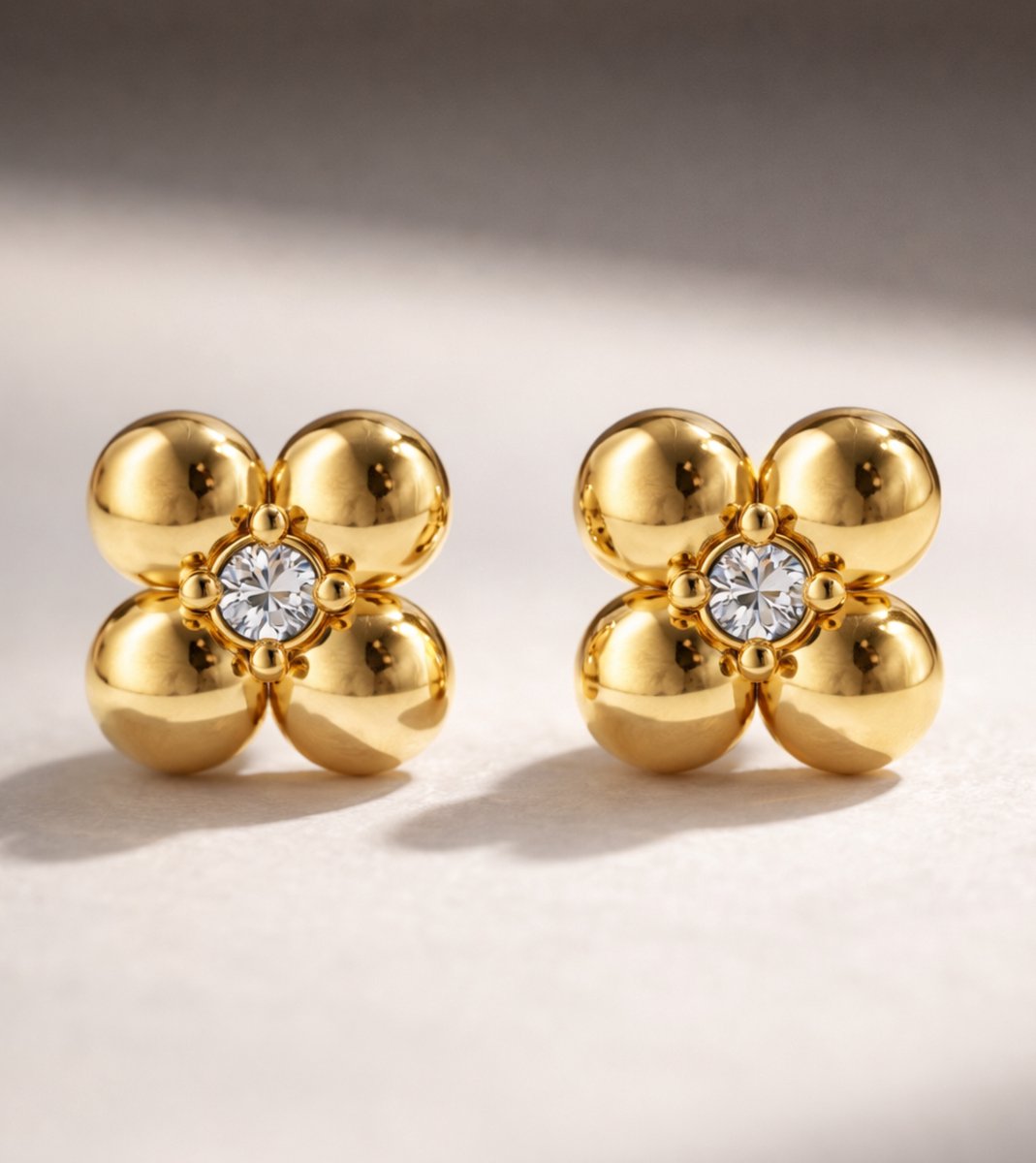 Lundholm Melle goudkleurige verfijnde oorknopjes voor dames met klein bloem ontwerp en zirkonia steen in het hart, gefotografeerd op crème ondergrond voor close-up productshot. Premium stainless steel met PVD gold plating van 2.5 micron, waterproof en hypoallergeen. Een subtiel cadeau voor vrouw, moeder of vriendin op Moederdag of verjaardag in Scandinavisch design.