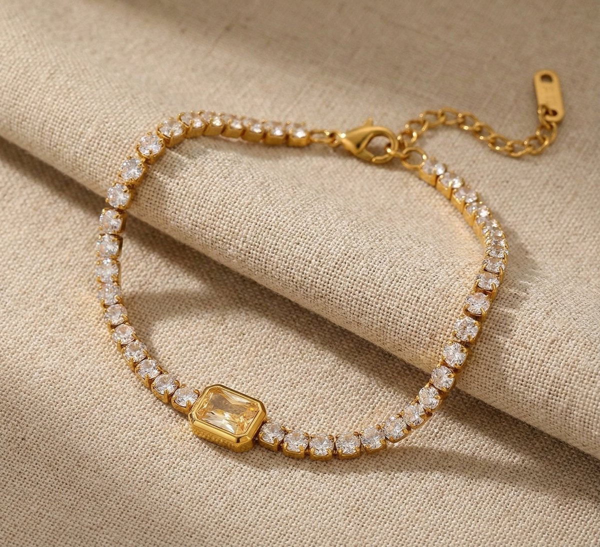 Lundholm My Jasmijn Serie luxe armband voor dames in goudkleurig met zirkonia stenen in tennis-stijl met centrale rechthoekige zirkonia op linnen achtergrond, gemaakt van premium stainless steel met PVD coating van 2.5 micron, waterproof en hypoallergeen, perfect cadeau voor vrouw, moederdag of verjaardag in Scandinavisch design.