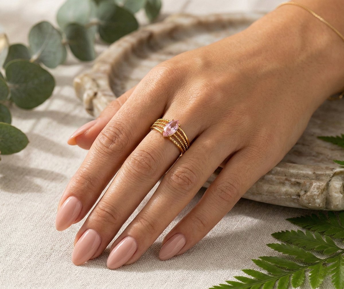 Lundholm My Juliette goudkleurige verstelbare cuff ring voor dames met grote roze natuursteen in vintage ontwerp, gedragen door vrouw met hand op linnen en rotan met eucalyptus en varen voor warme lookbook lifestyle shoot. Premium stainless steel met PVD gold plating van 2.5 micron, waterproof en hypoallergeen. Een romantisch cadeau voor vrouw, moeder of vriendin op Moederdag of verjaardag in Scandinavisch design.