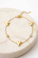 Lundholm My Jutta goudkleurige armband voor dames met romantische hartjes en fonkelende zirkonia steentjes, gefotografeerd op crème marmeren schaal voor warme productshot. Premium stainless steel met PVD gold plating van 2.5 micron, waterproof en hypoallergeen. Een romantisch cadeau voor vrouw, moeder of vriendin op Moederdag of verjaardag in Scandinavisch design.