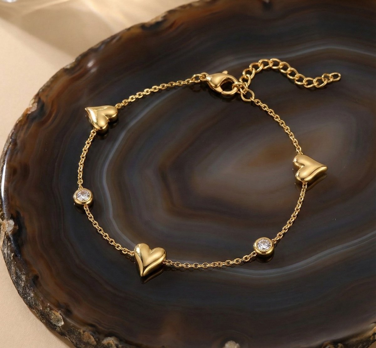 Lundholm My Jutta goudkleurige armband voor dames met hartjes en fonkelende zirkonia steentjes, gefotografeerd op donkere agaat natuursteen schaal voor sfeervolle productshot. Premium stainless steel met PVD gold plating van 2.5 micron, waterproof en hypoallergeen. Een romantisch cadeau voor vrouw, moeder of vriendin op Moederdag of verjaardag in Scandinavisch design.