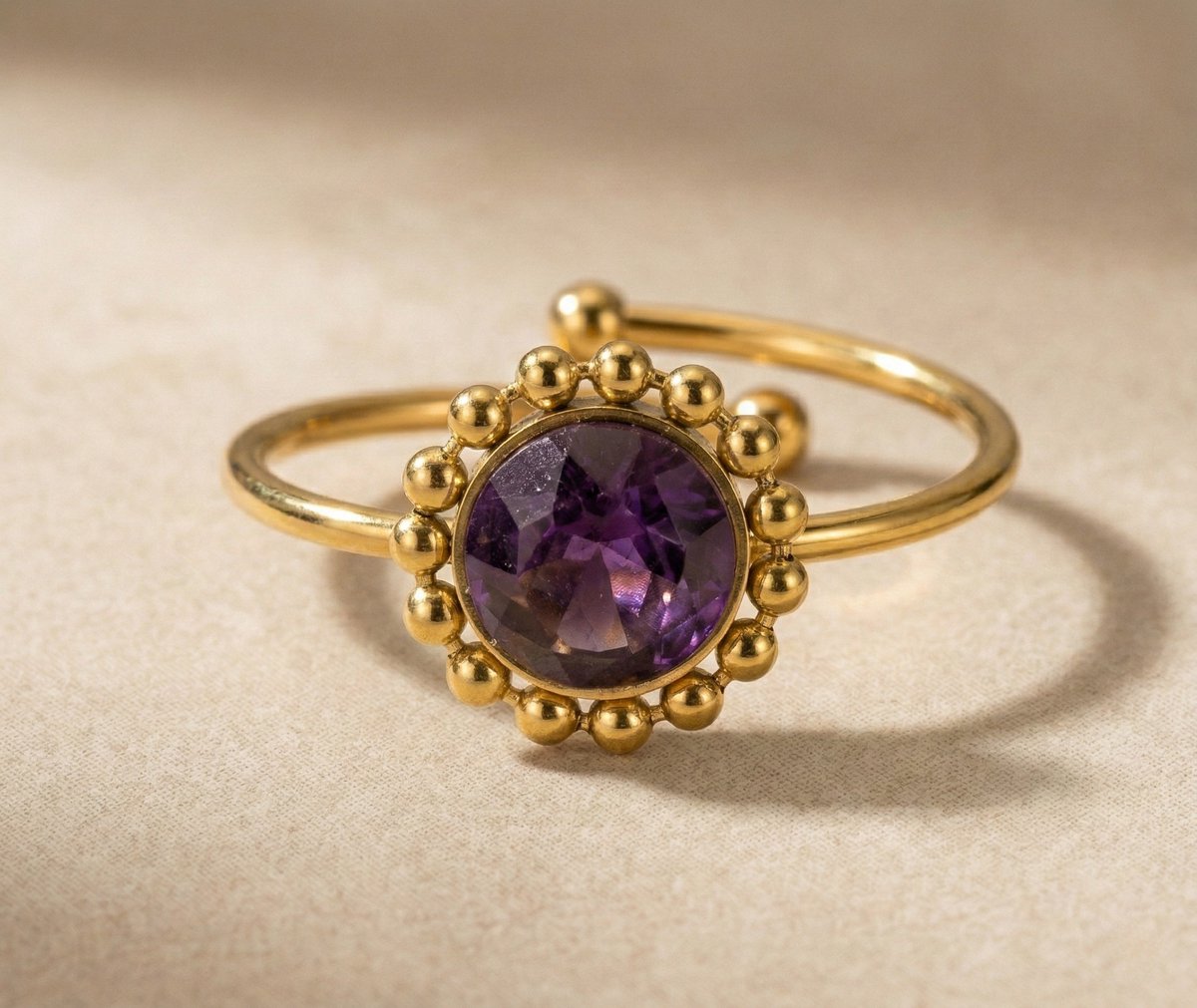Lundholm My Lilly goudkleurige verstelbare ring voor dames met paarse amethist natuursteen omringd door gouden bolletjes in vintage zonnebloem ontwerp, gefotografeerd op crème ondergrond voor warme productshot. Premium stainless steel met PVD gold plating van 2.5 micron, waterproof en hypoallergeen. Een romantisch cadeau voor vrouw, moeder of vriendin op Moederdag of verjaardag in Scandinavisch design.