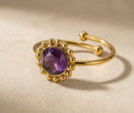 Lundholm My Lilly goudkleurige verstelbare ring voor dames met paarse amethist natuursteen omringd door gouden bolletjes in vintage zonnebloem ontwerp, gefotografeerd op crème stof voor warme productshot. Premium stainless steel met PVD gold plating van 2.5 micron, waterproof en hypoallergeen. Een romantisch cadeau voor vrouw, moeder of vriendin op Moederdag of verjaardag in Scandinavisch design.