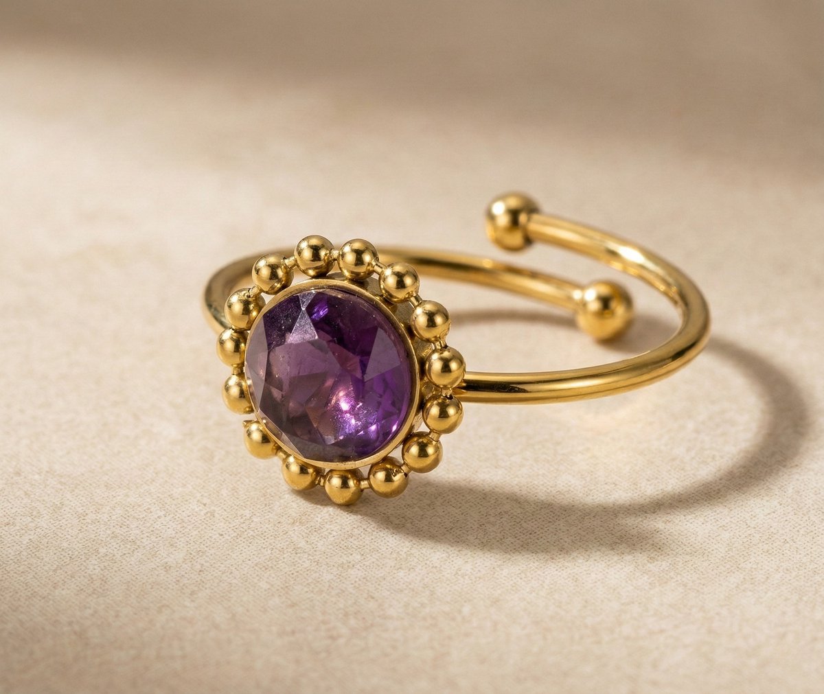 Lundholm My Lilly goudkleurige verstelbare ring voor dames met paarse amethist natuursteen omringd door gouden bolletjes in vintage zonnebloem ontwerp, gefotografeerd op crème stof voor warme productshot. Premium stainless steel met PVD gold plating van 2.5 micron, waterproof en hypoallergeen. Een romantisch cadeau voor vrouw, moeder of vriendin op Moederdag of verjaardag in Scandinavisch design.