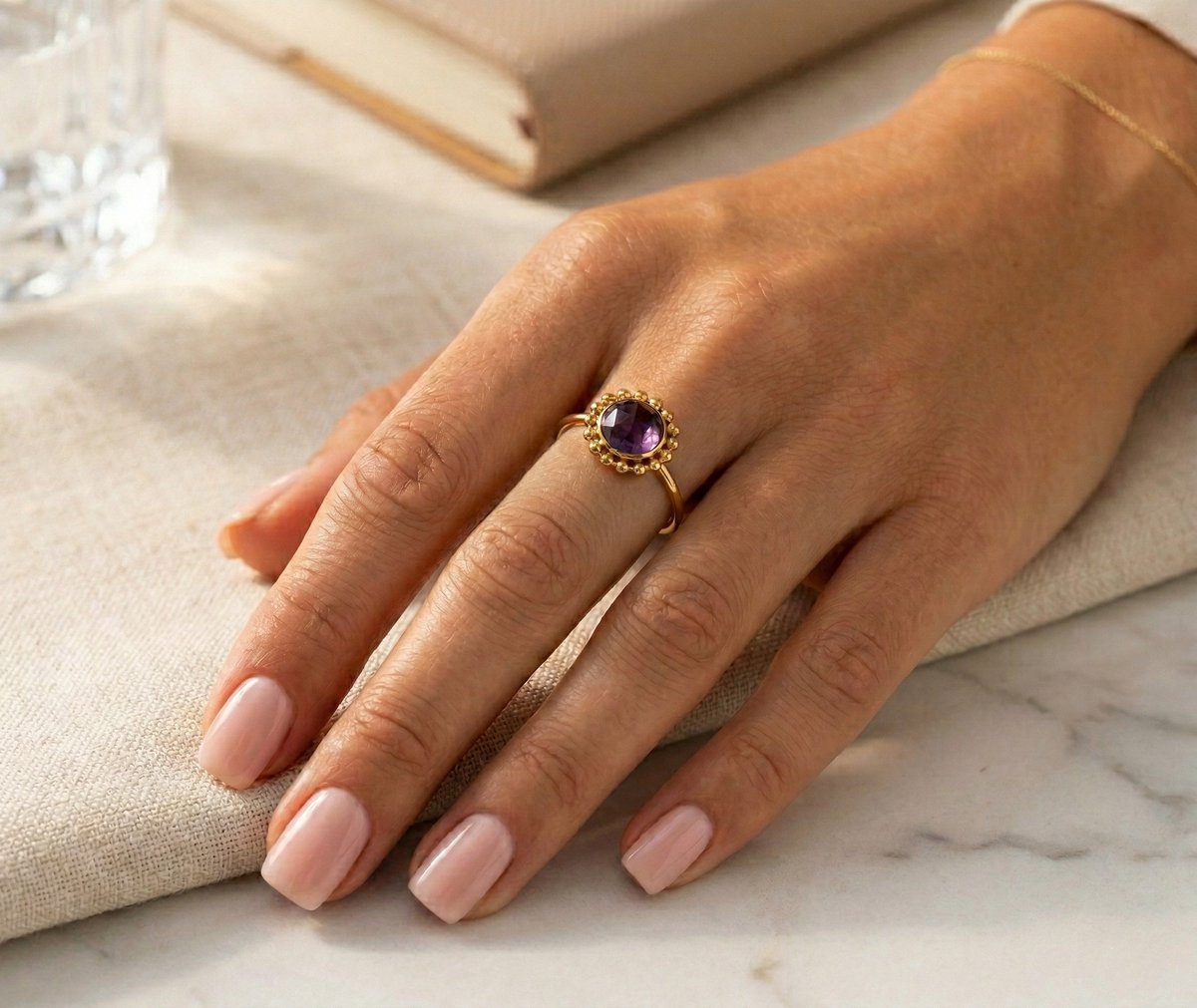 Lundholm My Lilly goudkleurige verstelbare ring voor dames met paarse amethist natuursteen in vintage zonnebloem ontwerp, gedragen door vrouw met hand op linnen tafelloper bij boek en kristal glas voor verfijnde lookbook lifestyle shoot. Premium stainless steel met PVD gold plating van 2.5 micron, waterproof en hypoallergeen. Een romantisch cadeau voor vrouw, moeder of vriendin op Moederdag of verjaardag in Scandinavisch design.