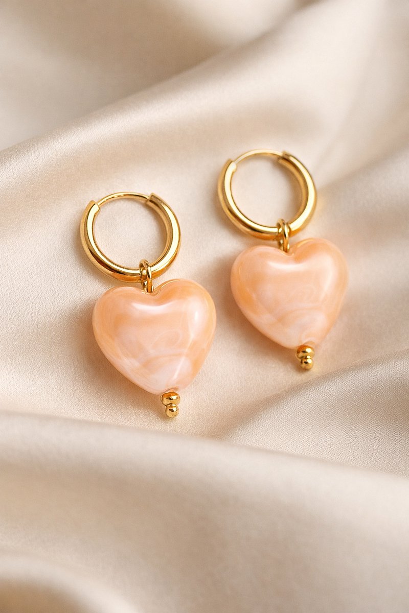 Lundholm My Merle Serie luxe oorbellen voor dames in goudkleurig met beige hartjes hanger op zijde achtergrond, gemaakt van premium stainless steel met PVD coating van 2.5 micron, waterproof en hypoallergeen, perfect cadeau voor vrouw, moederdag of verjaardag in Scandinavisch design.