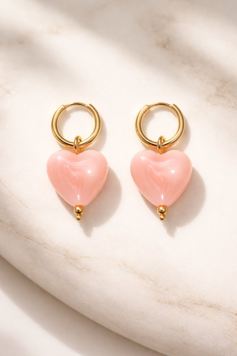 Lundholm My Merle Serie luxe oorbellen voor dames in goudkleurig met roze hartjes hanger op marmeren ondergrond, gemaakt van premium stainless steel met PVD coating van 2.5 micron, waterproof en hypoallergeen, perfect cadeau voor vrouw, moederdag of verjaardag in Scandinavisch design.