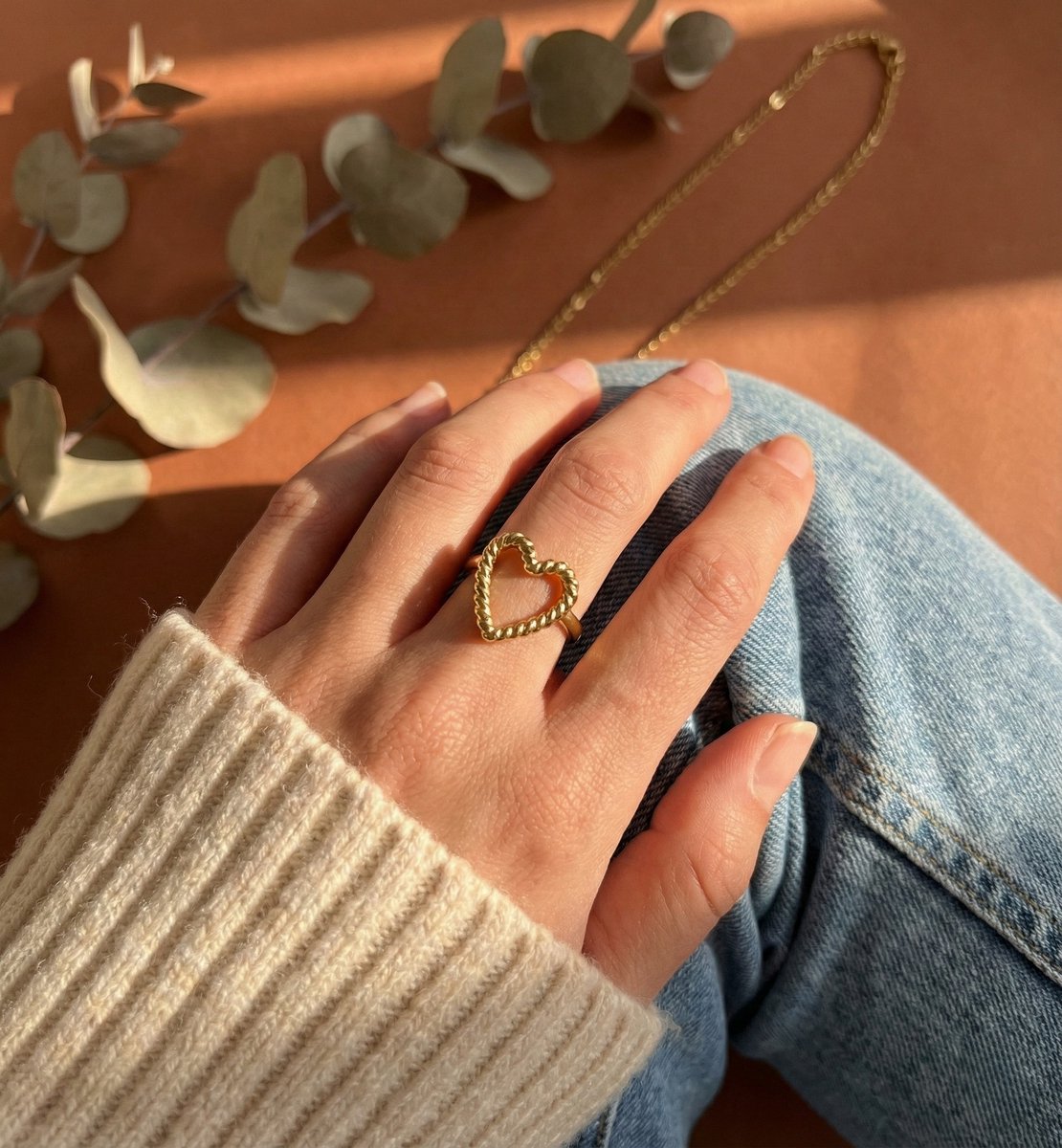 Lundholm My Rae goudkleurige verstelbare statement ring voor dames met open hart ontwerp en getwist textuur, gedragen door vrouw met crème trui en denim voor warme lookbook lifestyle shoot met eucalyptus. Premium stainless steel met PVD gold plating van 2.5 micron, waterproof en hypoallergeen. Een romantisch cadeau voor vrouw, moeder of vriendin op Moederdag of verjaardag in Scandinavisch design.