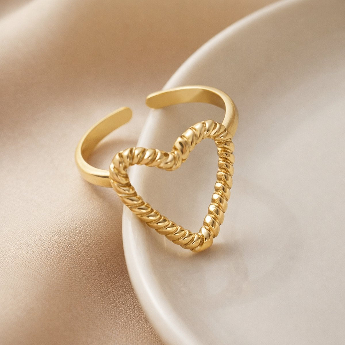 Lundholm My Rae goudkleurige verstelbare statement ring voor dames met open hart ontwerp en getwiste touw textuur, gefotografeerd op witte keramische schaal voor warme productshot. Premium stainless steel met PVD gold plating van 2.5 micron, waterproof en hypoallergeen. Een romantisch cadeau voor vrouw, moeder of vriendin op Moederdag of verjaardag in Scandinavisch design.
