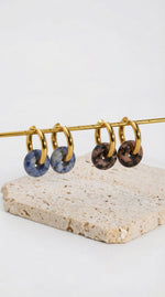 Lundholm My Sally Serie luxe oorbellen voor dames in goudkleurig, twee varianten met blauwe en bruine natuursteen donut hanger op travertijn display, gemaakt van premium stainless steel met PVD coating van 2.5 micron, waterproof en hypoallergeen, perfect cadeau voor vrouw, moederdag of verjaardag in Scandinavisch design.