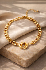Lundholm My Tilda goudkleurige schakelarmband voor dames van 16,4 cm met verbonden ringen detail, gefotografeerd op crème stof en travertijn steen voor warme productshot. Premium stainless steel met PVD gold plating van 2.5 micron, waterproof en hypoallergeen. Een tijdloos cadeau voor vrouw, moeder of vriendin op Moederdag of verjaardag in Scandinavisch design.