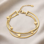 Lundholm My Yaar goudkleurige meerdere lagen schakelarmband voor dames van 16,4 cm met snake chain en hartjes accenten, gefotografeerd op crème zijde voor elegante productshot. Premium stainless steel met PVD gold plating van 2.5 micron, waterproof en hypoallergeen. Een romantisch cadeau voor vrouw, moeder of vriendin op Moederdag of verjaardag in Scandinavisch design.