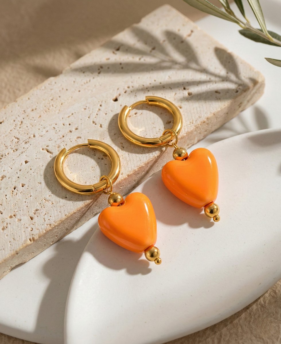 Lundholm Neeltje goudkleurige huggie oorbellen voor dames met grote oranje hartjes hangers, gefotografeerd op travertijn natuursteen met olijfblad voor warme productshot. Perfect voor Koningsdag accessoires. Premium stainless steel met PVD gold plating van 2.5 micron, waterproof en hypoallergeen. Een vrolijk cadeau voor vrouw, moeder of vriendin op Koningsdag, Moederdag of verjaardag in Scandinavisch design.