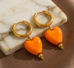 Lundholm Neeltje goudkleurige huggie oorbellen voor dames met grote oranje hartjes hangers, gefotografeerd op witte marmeren ondergrond voor warme productshot. Perfect voor Koningsdag accessoires. Premium stainless steel met PVD gold plating van 2.5 micron, waterproof en hypoallergeen. Een vrolijk cadeau voor vrouw, moeder of vriendin op Koningsdag, Moederdag of verjaardag in Scandinavisch design.