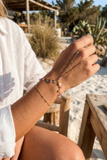 Lundholm Saar goudkleurig handsieraad voor dames met hand chain ontwerp dat armband en ring verbindt, voorzien van kleurrijke bedels, gedragen door vrouw outdoor op het strand voor warme zomerse lookbook lifestyle shoot. Premium stainless steel met PVD gold plating van 2.5 micron, waterproof en hypoallergeen. Een uniek cadeau voor vrouw, moeder of vriendin op Moederdag of verjaardag in Scandinavisch design.