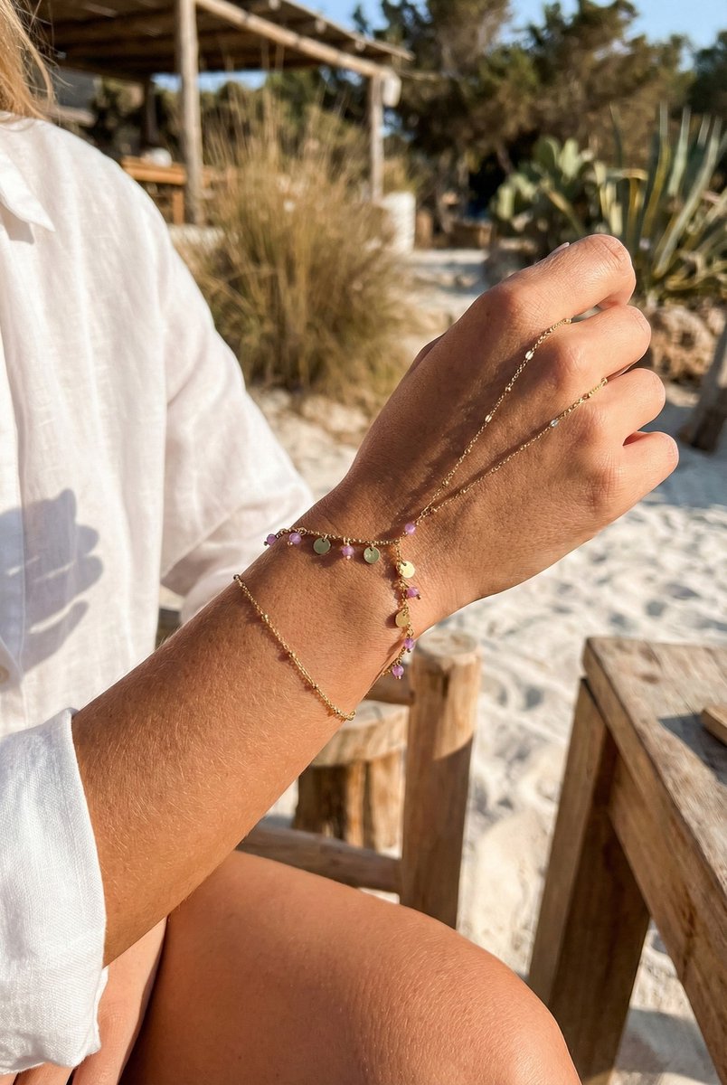 Lundholm Saar goudkleurig handsieraad voor dames met hand chain ontwerp dat armband en ring verbindt, voorzien van kleurrijke bedels, gedragen door vrouw outdoor op het strand voor warme zomerse lookbook lifestyle shoot. Premium stainless steel met PVD gold plating van 2.5 micron, waterproof en hypoallergeen. Een uniek cadeau voor vrouw, moeder of vriendin op Moederdag of verjaardag in Scandinavisch design.