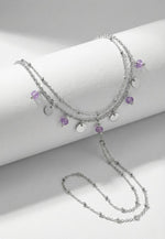Lundholm Saar zilverkleurig handsieraad voor dames met hand chain ontwerp dat armband en ring verbindt, voorzien van paarse amethist en zilveren bedels, gefotografeerd op witte ondergrond voor stijlvolle productshot. Premium stainless steel met PVD silver plating, waterproof en hypoallergeen. Een uniek cadeau voor vrouw, moeder of vriendin op Moederdag of verjaardag in Scandinavisch design.