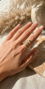 Lundholm Waves Serie luxe ring voor dames in goudkleurig met drie laags golvend design verstelbaar gedragen aan hand van model in lifestyle setting met pampasgras, gemaakt van premium stainless steel met PVD coating van 2.5 micron, waterproof en hypoallergeen, perfect cadeau voor vrouw, moederdag of verjaardag in Scandinavisch design.