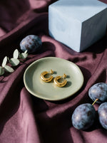 Lundholm Sol chunky hoop oorknopjes goudkleurig op een saliegroen schaaltje met pruimen en plum satijn - premium stainless steel PVD gold plated, Scandinavisch design