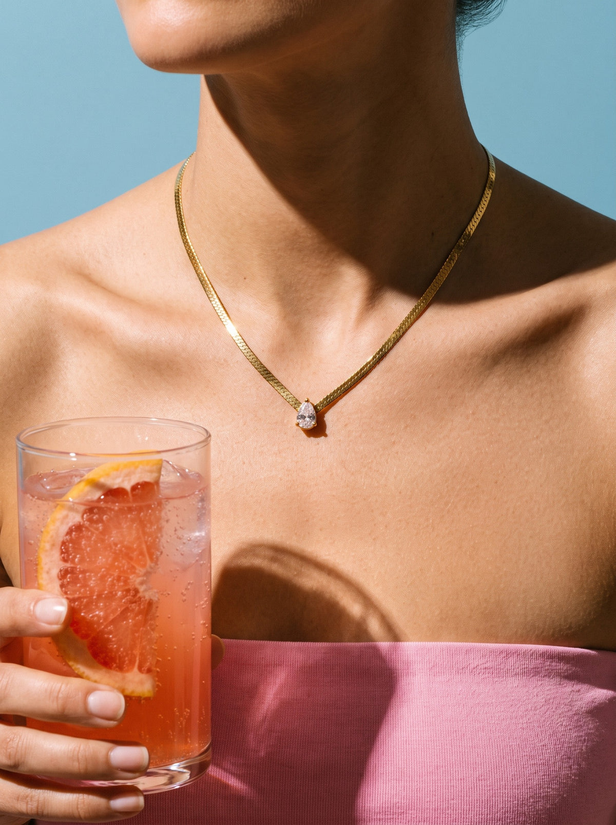 Vrouw draagt de Lundholm Ariane platte schakelketting met zirkonia goudkleurig, met een glas grapefruit soda
