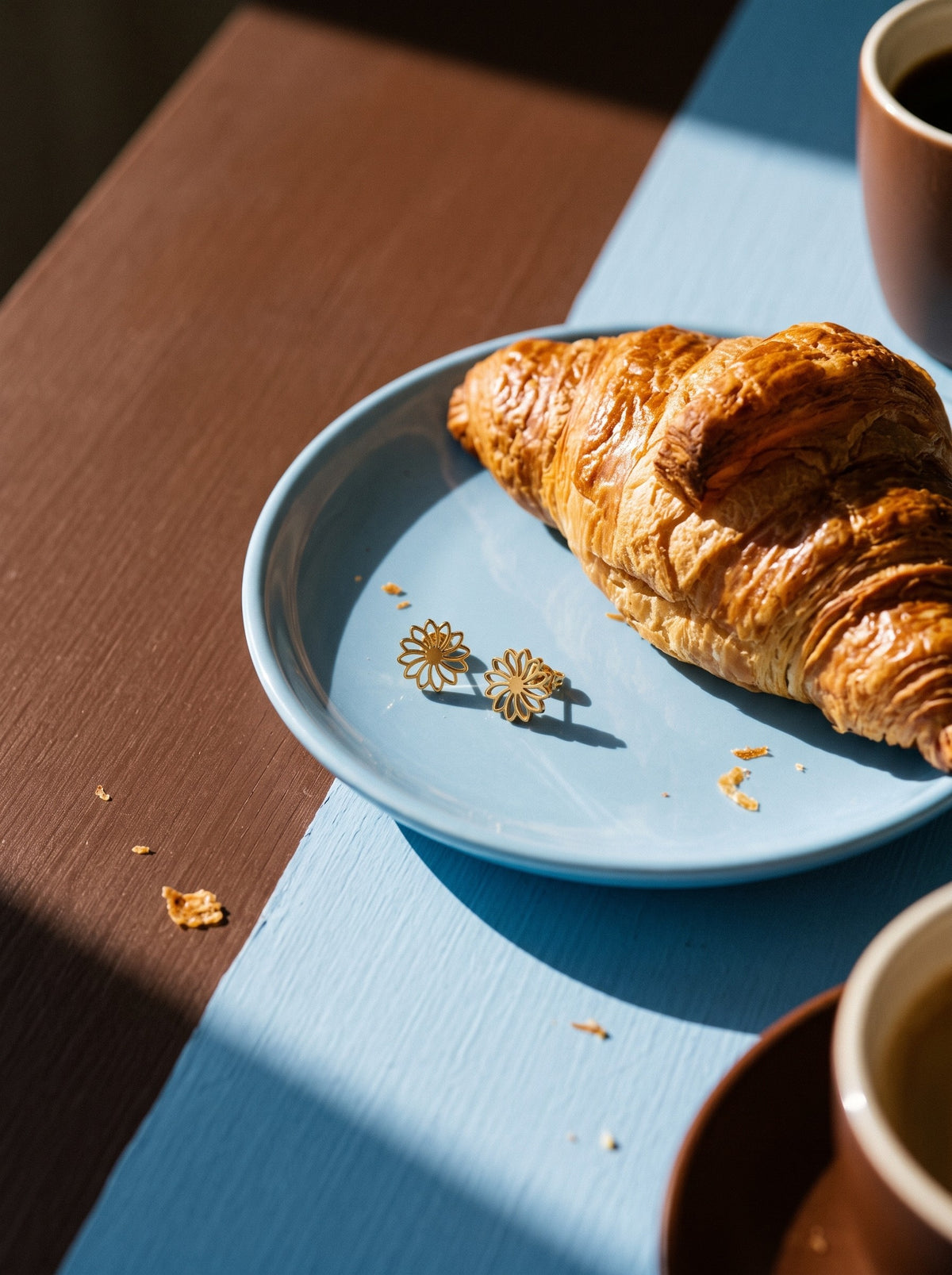 Lundholm Aya bloem oorknopjes goudkleurig op een blauw bordje naast een croissant