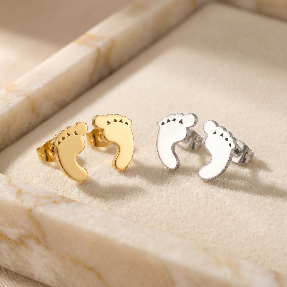 Lundholm Bebe babyvoetjes oorknopjes in goud en zilver naast elkaar op marmer
