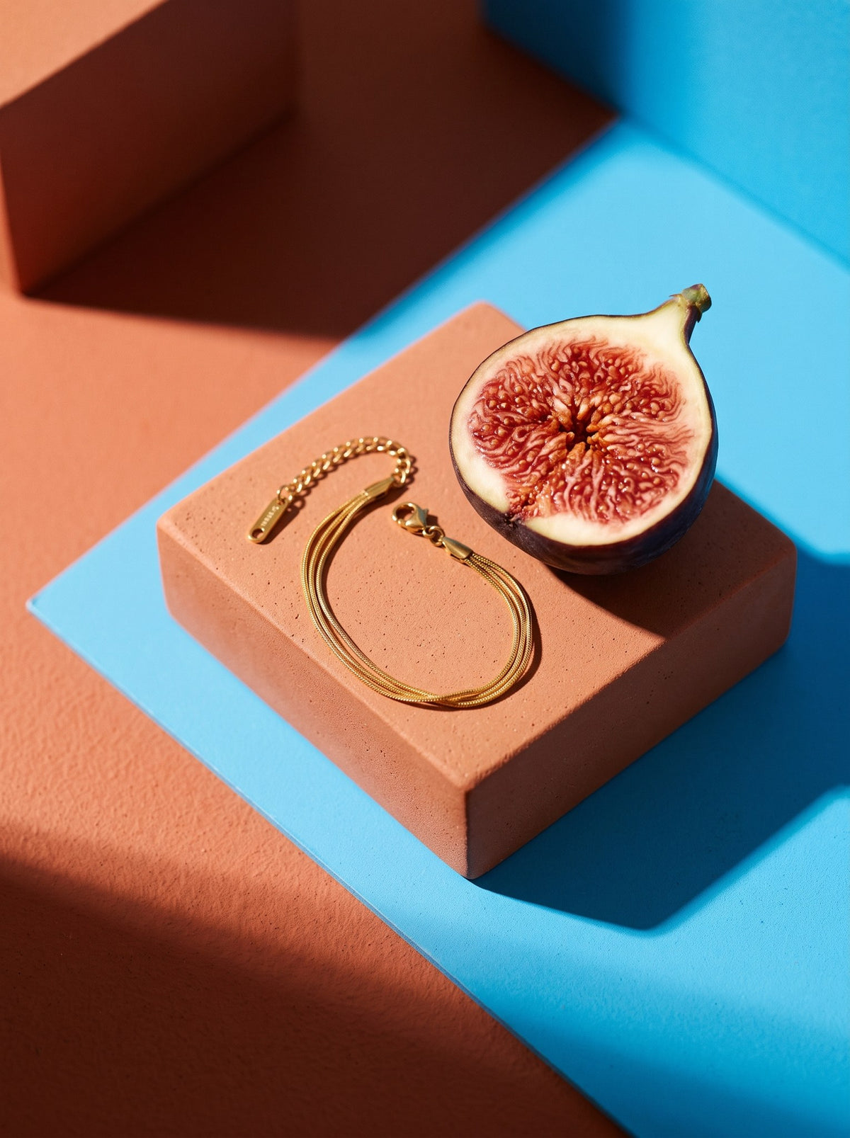 Lundholm Bibi drielaagse armband goudkleurig op een terracotta blok naast een halve vijg